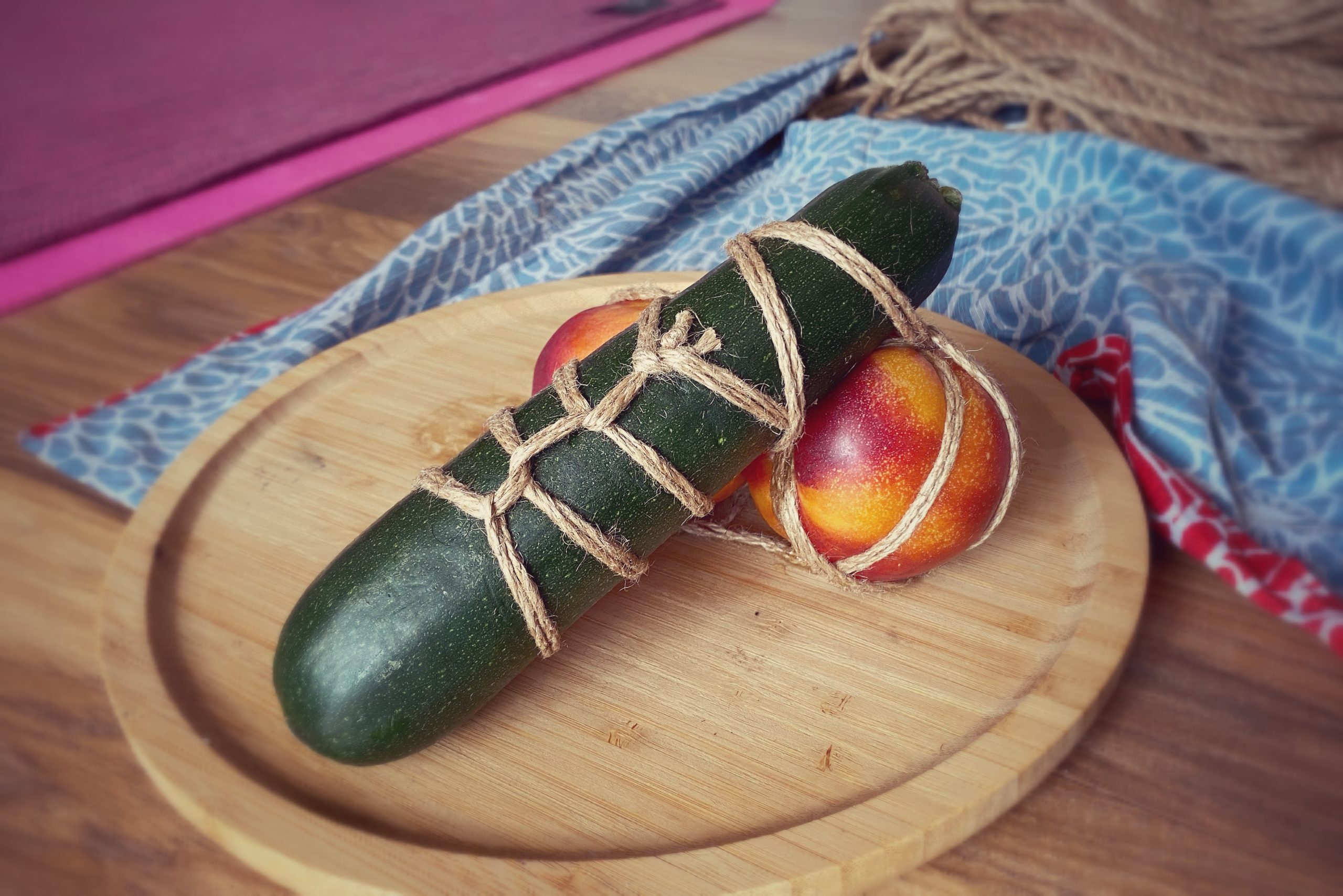 a wooden plate with a zucchini and two peaches which are tied together looking like a penis and balls