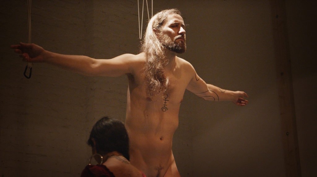 Portrait of a person with long hair and a beard in a ritual hook suspension setting.