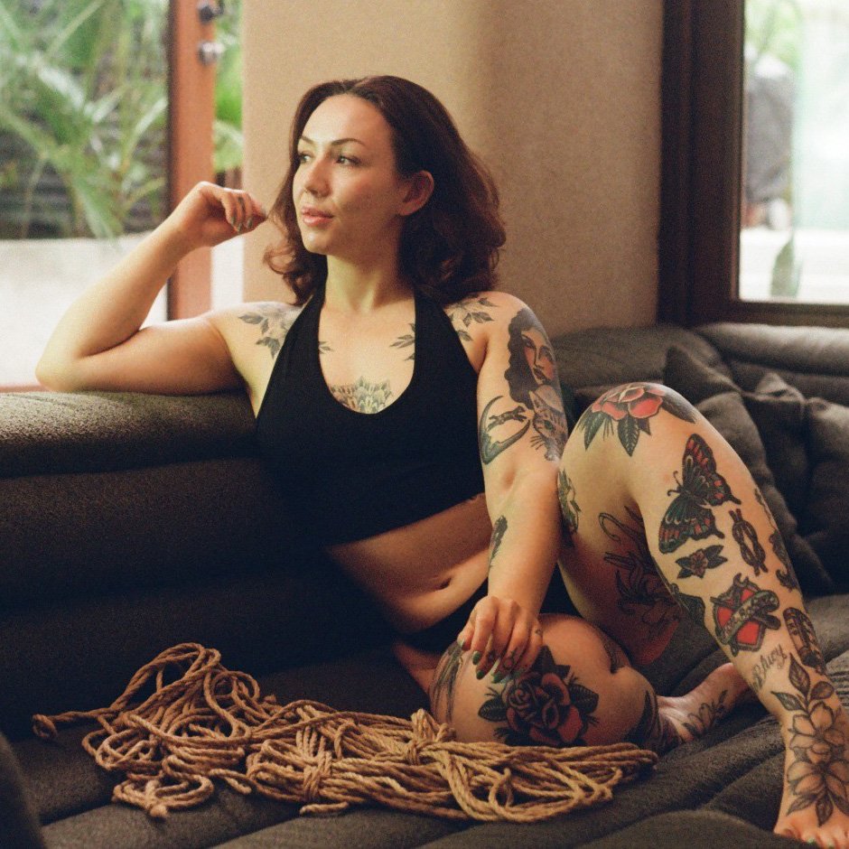 Tattooed woman sitting on sofa with rope nearby.