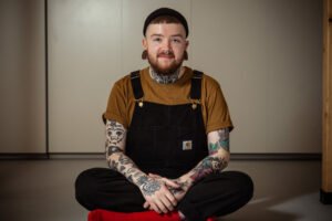 Tattooed man in overalls sitting cross-legged indoors